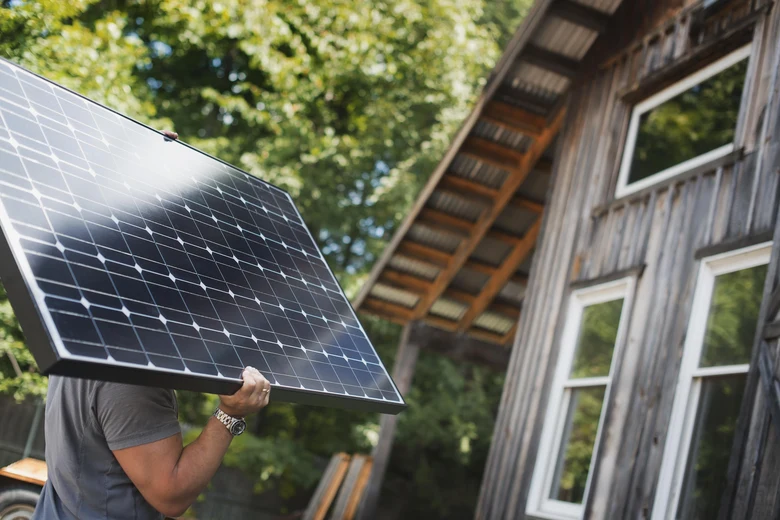 Muž nese solární panely k dřevěnému domu pro využití sluneční energie.