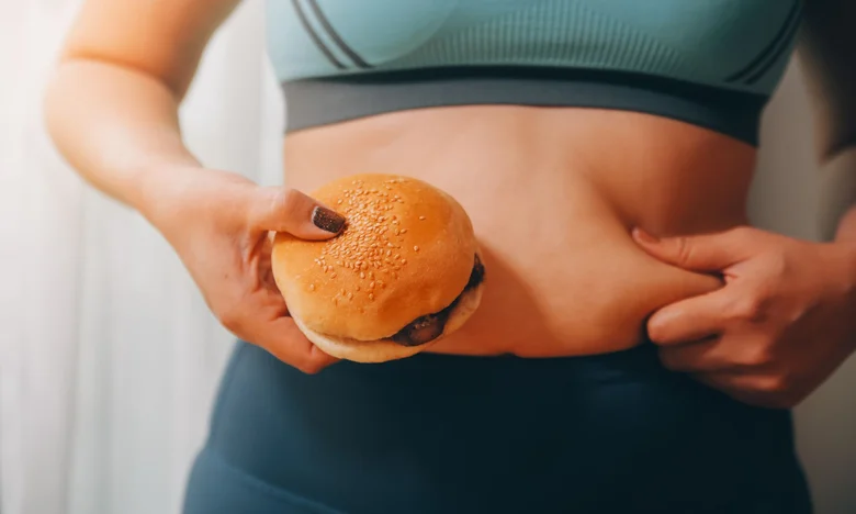 Žena drží hamburger a táhne si břišní tuk.