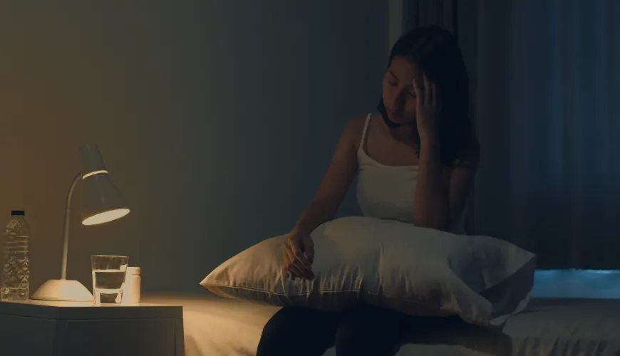 Žena probuzená v noci, drží si hlavu, vedle postele stojí lampa a sklenice vody
