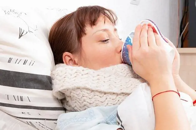Žena v posteli s šálou pije z hrnku, jako by měla nachlazení