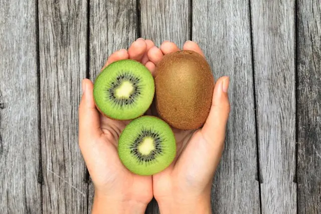 Dvě ruce drží celý i nakrájené zelené kiwi na dřevěném pozadí.