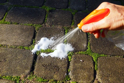 Ruka postřikuje plevel mezi kostkami pěnou z oranžové lahvičky.
