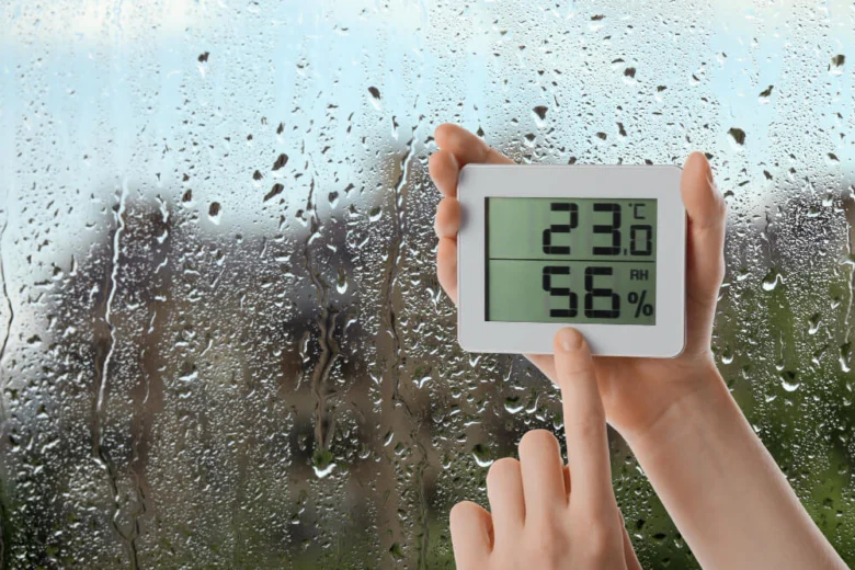 Osoba drží digitální vlhkoměr s teplotou 23°C a vlhkostí 56 % před rozmazaným oknem