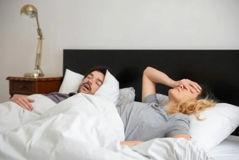 Muž chrápá v posteli, žena si zakrývá uši a tvář