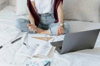 Student studuje na posteli s knihami a notebookem.