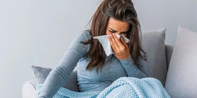 Žena sedí na gauči s rouškou v ruce při chřipce.