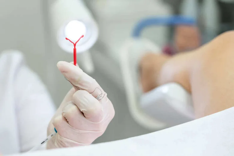 Rukou v rukavicích je drženo červené nitroděložní tělísko s krevními stopami.