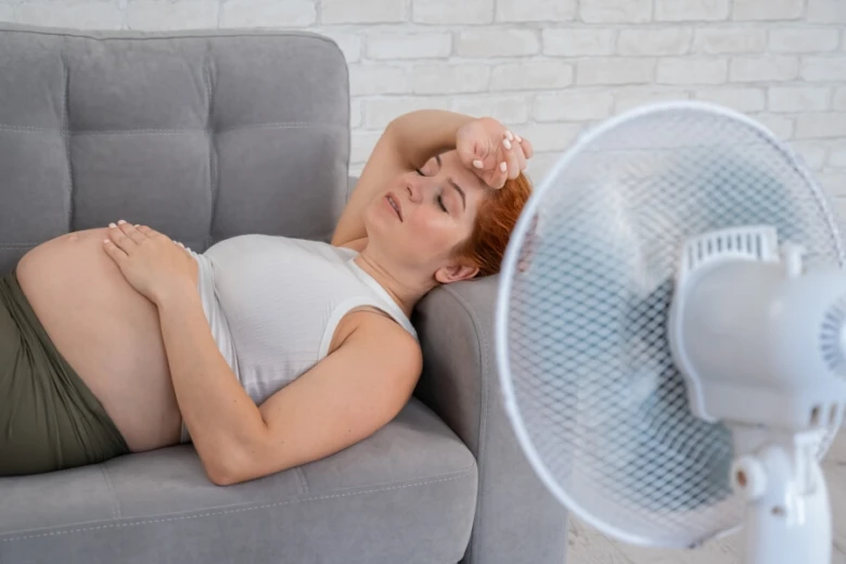 Těhotná žena leží na gauči s ventilátorem kvůli pocení.
