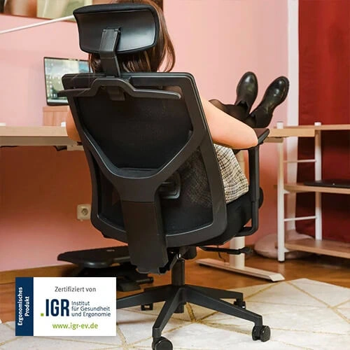 Člověk sedí v ergonomickém křesle u počítače s nohama na opěradlu.