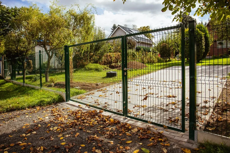 Zelený vyplétaný plot s bránou v zahradě pod stromy.